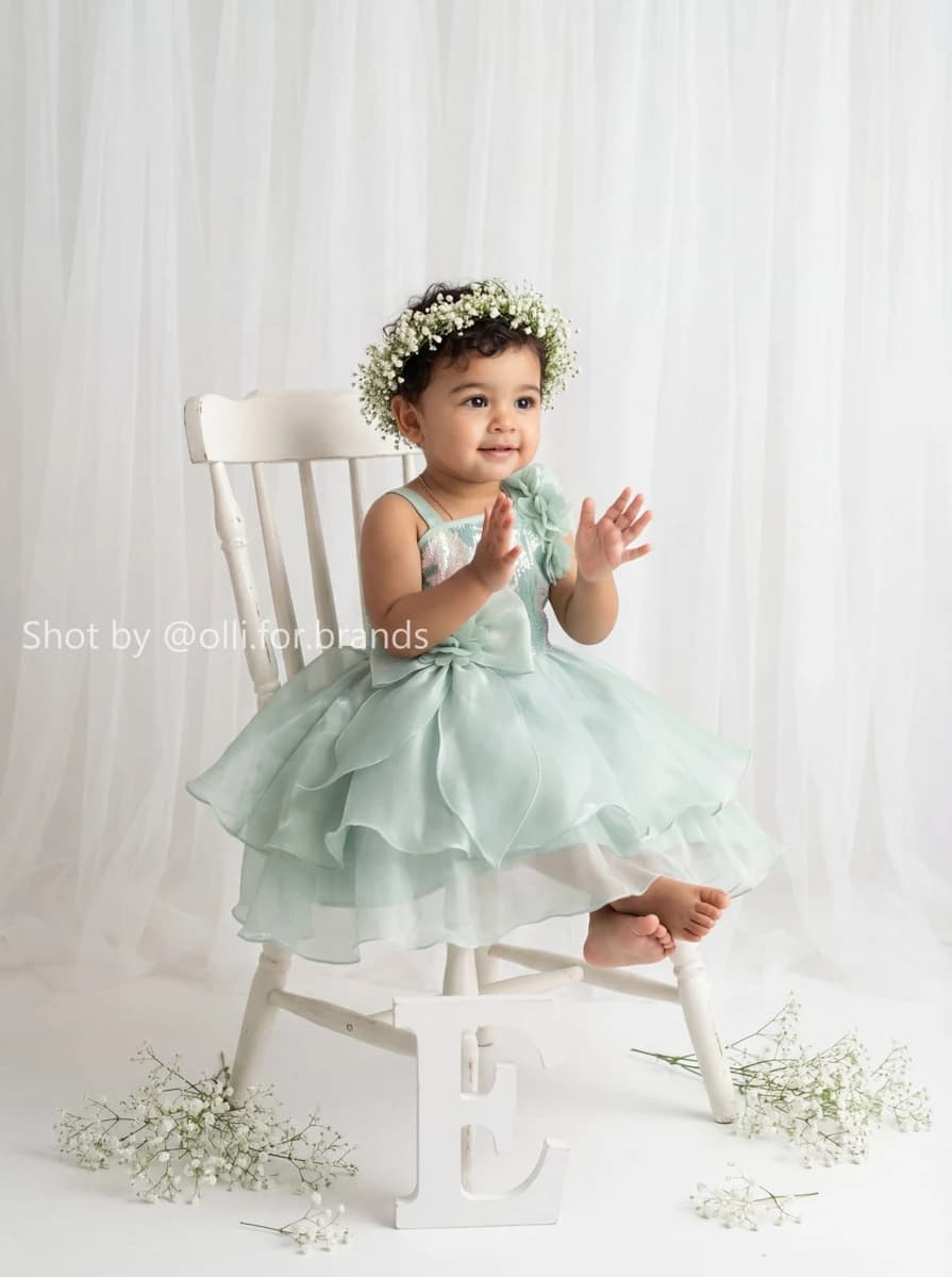 Toddler girl wearing a mint tulle dress sitting on a white chair with a floral crown.