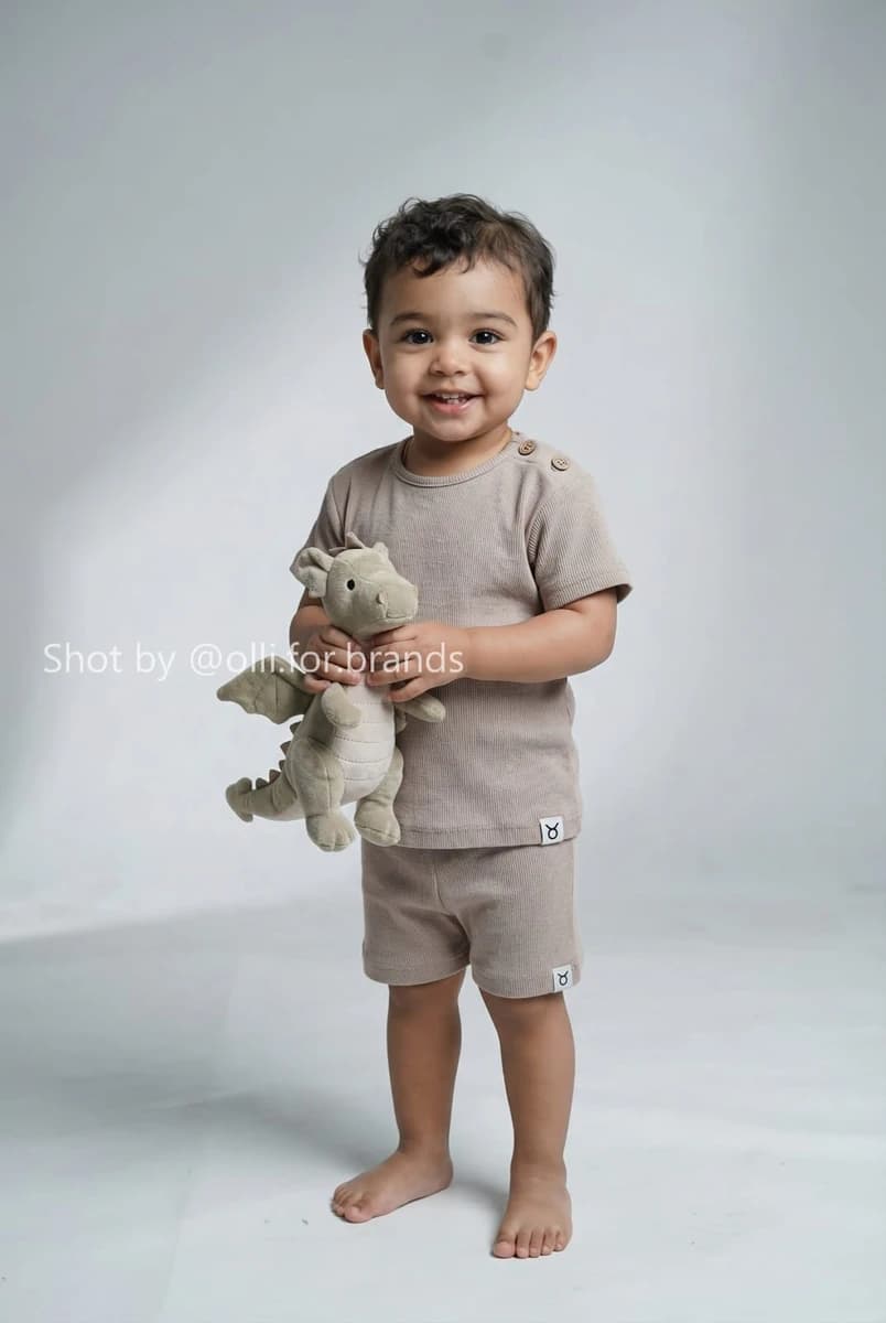 Smiling toddler wearing a ribbed short-sleeve top and matching shorts, holding a small plush dragon in a studio.