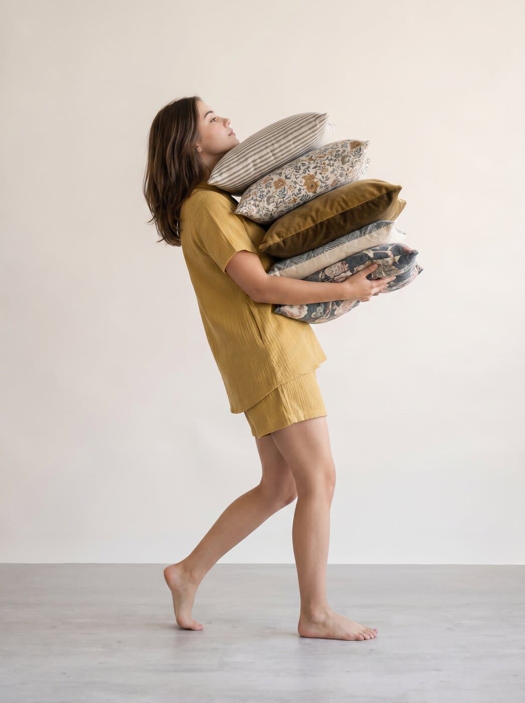 Woman in a mustard short-sleeve top and shorts lounge set carrying a stack of patterned cushions against a cream wall, an Olli catalog shot
