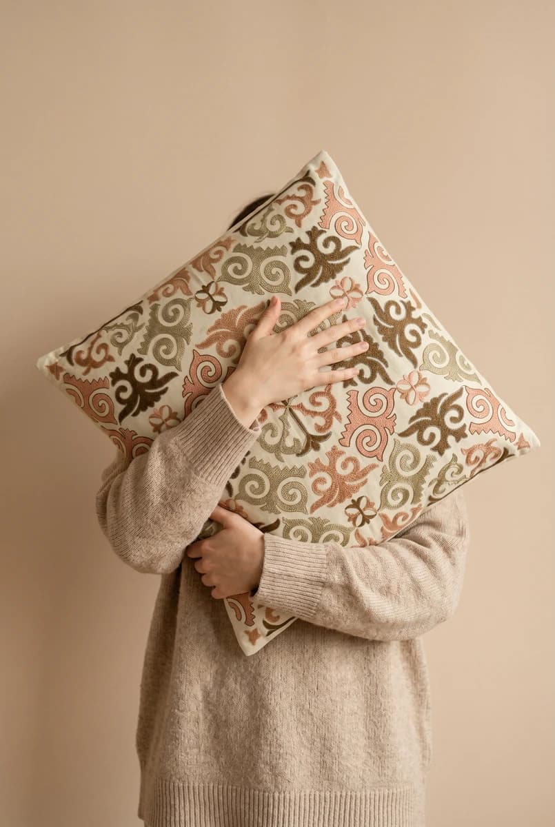 A person in a beige knit sweater holding a patterned square cushion in front of their face against a plain background.